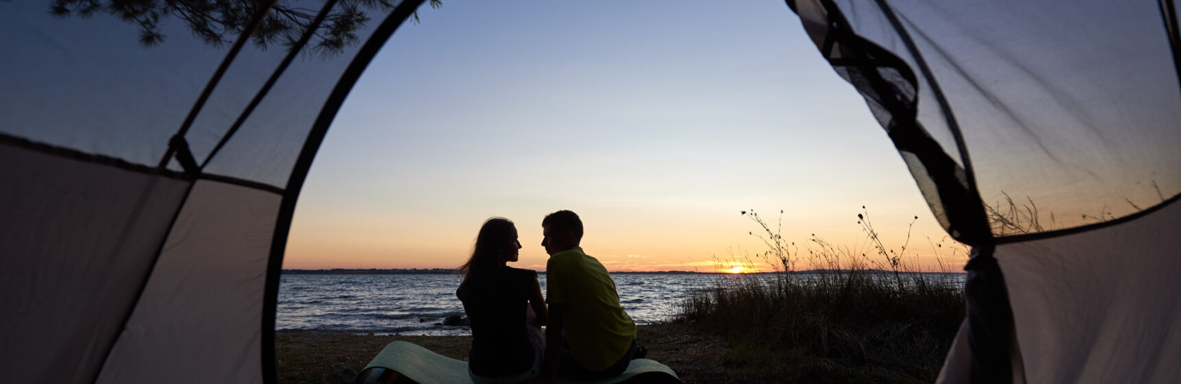 View from inside tourist tent. Silhouettes of young tourist romantic couple, man and woman sitting on lake shore on blue evening sky and crystal blue clear lake water background.