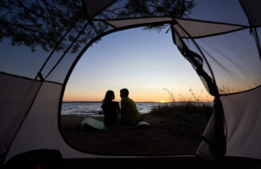 View from inside tourist tent. Silhouettes of young tourist romantic couple, man and woman sitting on lake shore on blue evening sky and crystal blue clear lake water background.