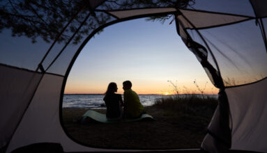 View from inside tourist tent. Silhouettes of young tourist romantic couple, man and woman sitting on lake shore on blue evening sky and crystal blue clear lake water background.